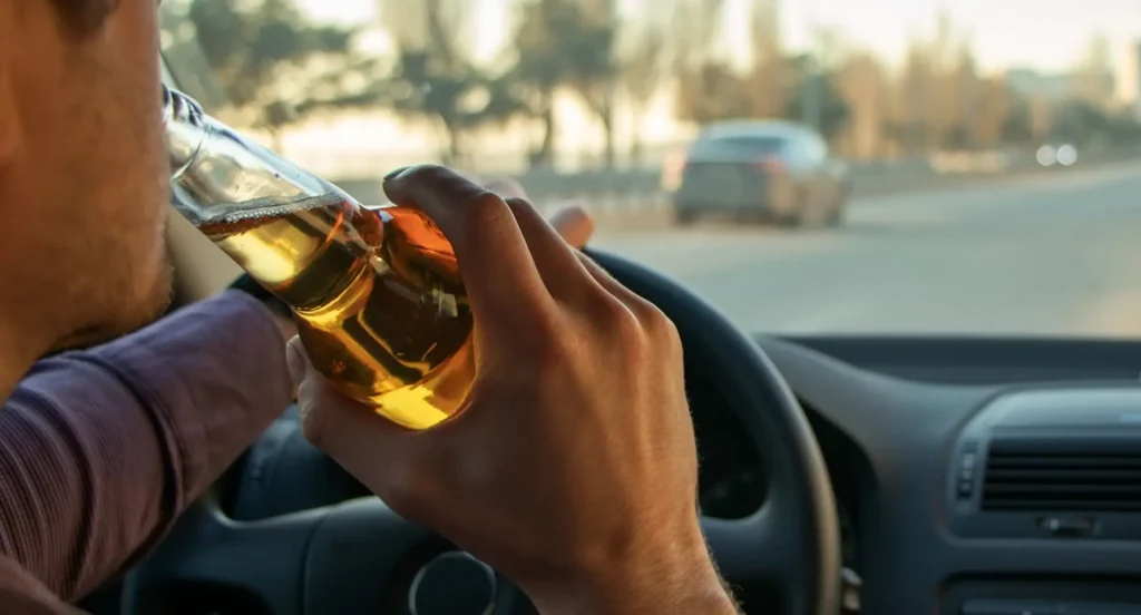 Man drinking alcohol while driving, holding bottle with both hands on the steering wheel.
