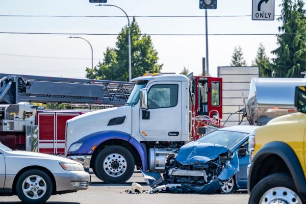 Crashed blue car with front-end damage at busy intersection near semi-truck and emergency responders.