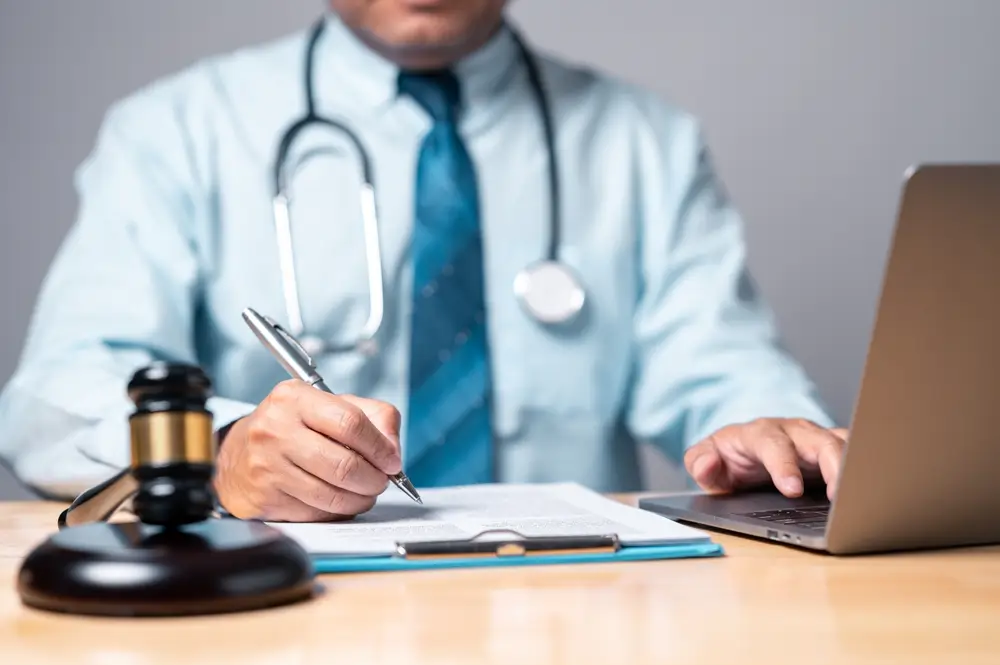 Doctor with stethoscope writing medical-legal report near gavel and laptop for personal injury case.