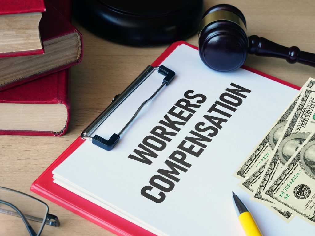 Clipboard labeled 'Workers Compensation' with money, gavel, and legal books nearby.