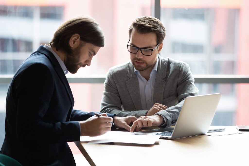 Two male professionals discussing documents in a modern office setting with a laptop on the desk.