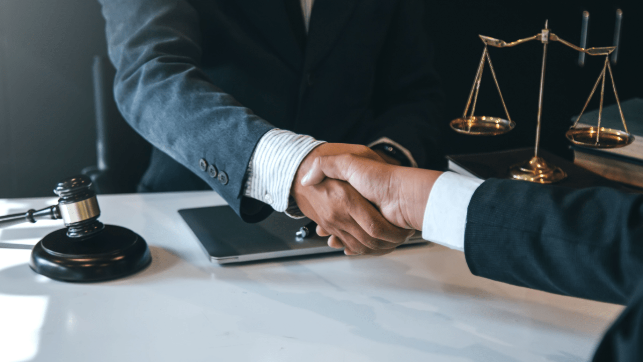 Two legal professionals shaking hands across a desk with a gavel, laptop, and justice scale — how to get new legal clients.