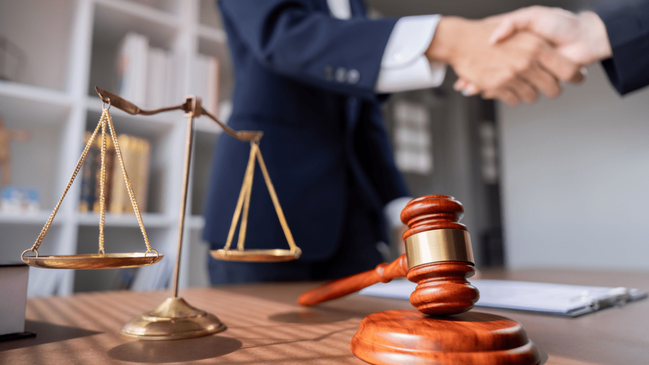 Close-up of legal symbols on a lawyer’s desk with two professionals shaking hands in the background, representing agreement and success in the legal field — is criminal defense profitable?